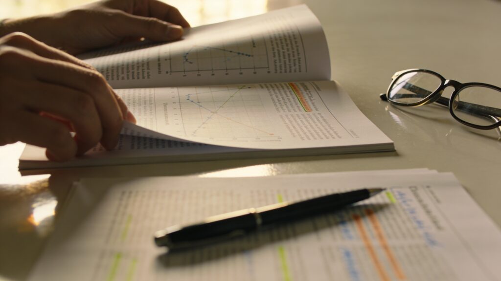 Main feuilletant un article scientifique imprimé avec des graphiques, un stylo posé sur une autre page annotée et une paire de lunettes sur la table, illustrant la lecture critique des publications.