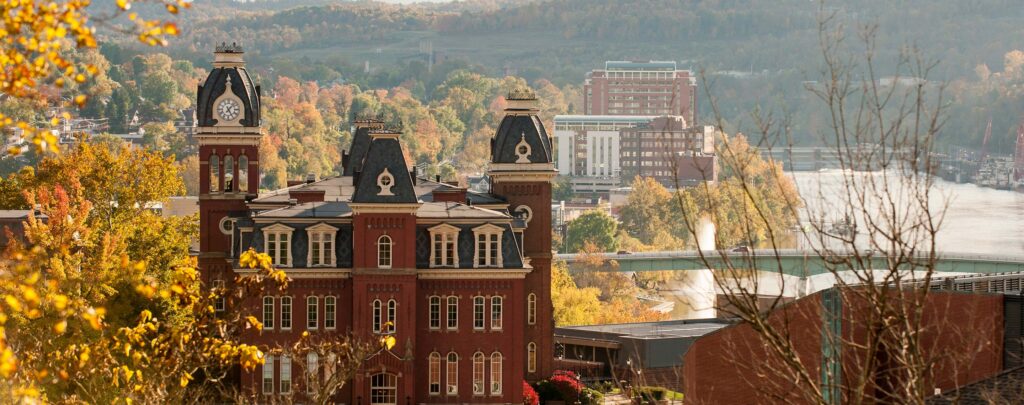 Vue panoramique du campus de West Virginia University à l’automne, avec un bâtiment historique en briques rouges au premier plan et la rivière en arrière‑plan.